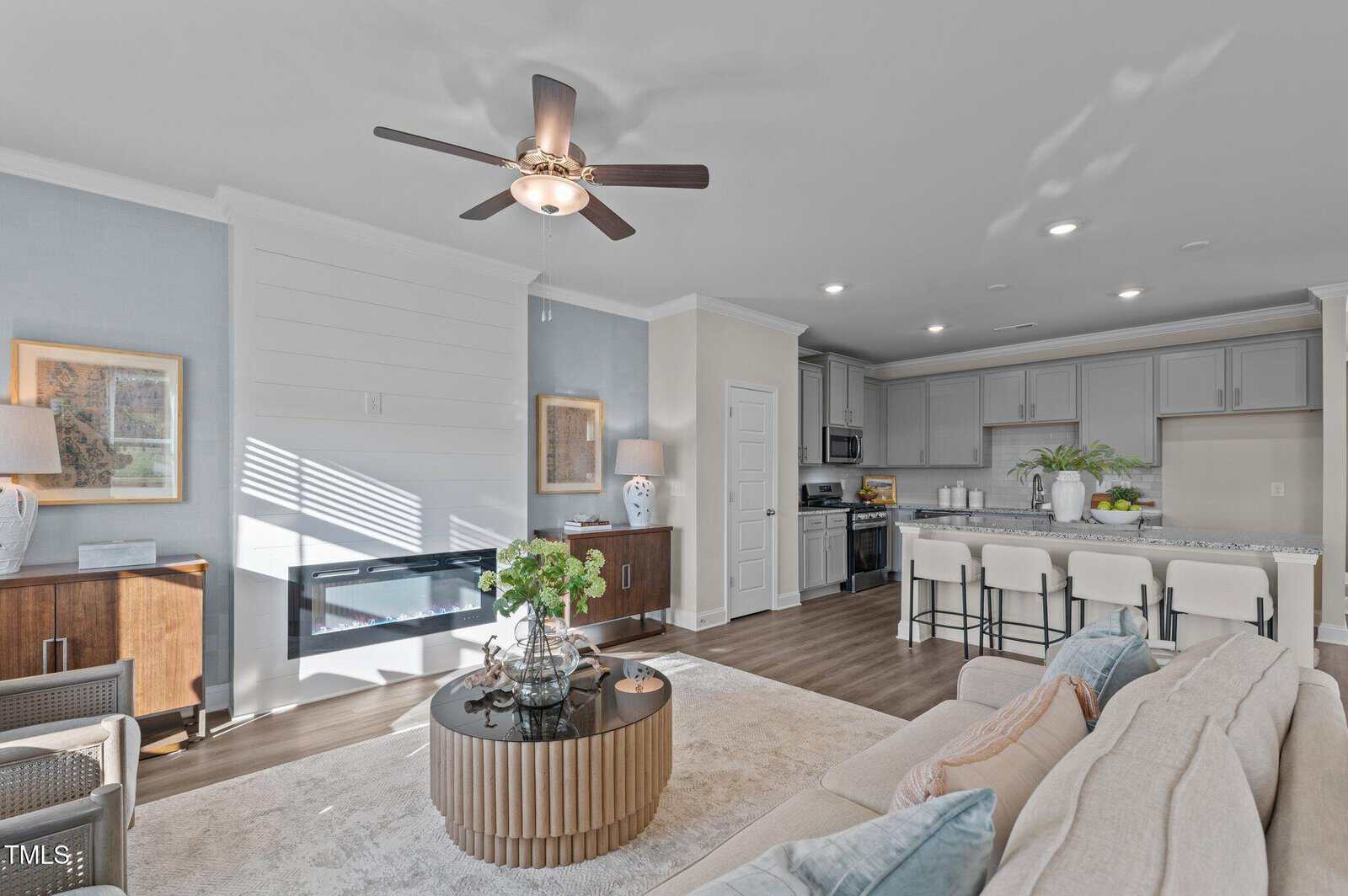 Bright, modern living room with beige sofa, round coffee table, wall-mounted TV, and open kitchen with bar seating. Ceiling fan and natural light enhance the airy atmosphere.