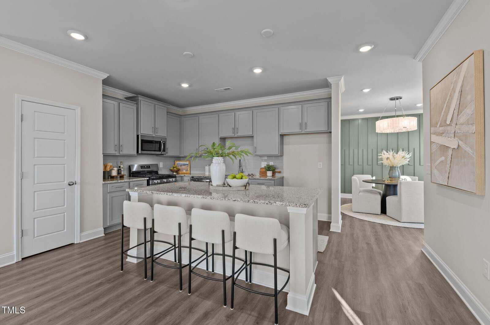 Modern kitchen with light gray cabinets, stainless steel appliances, a large granite island with four upholstered barstools, and decorative plants. The space opens to a dining area with a round table and green accent wall.