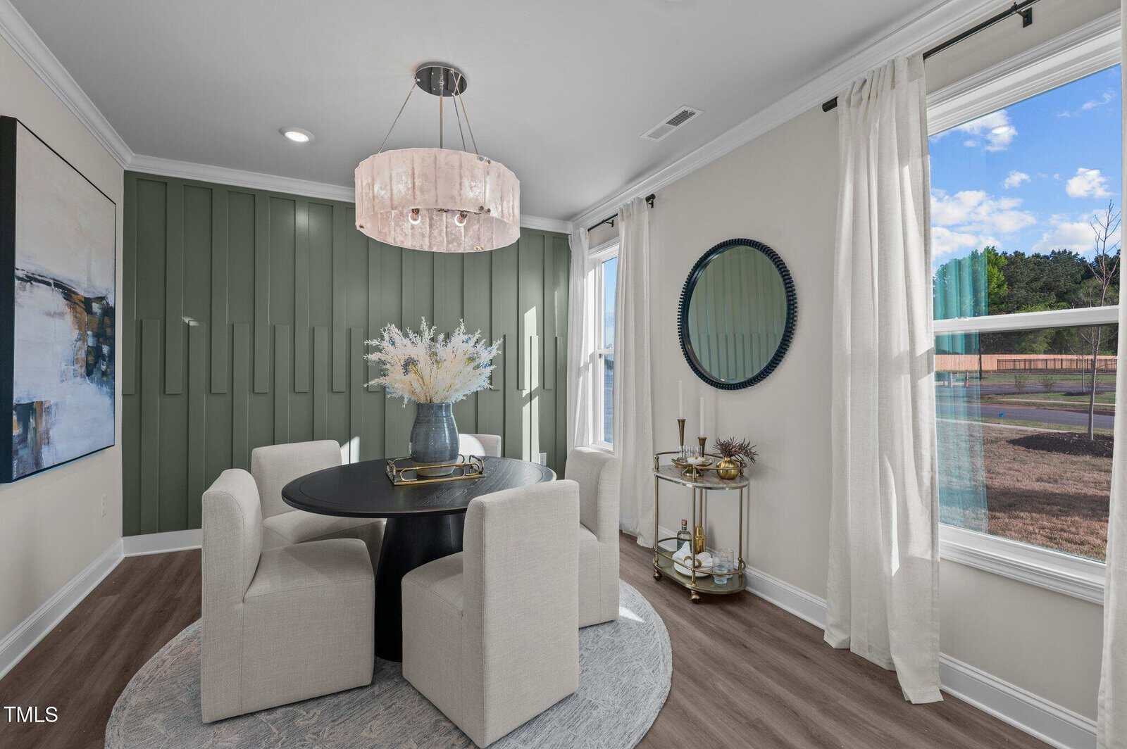 A modern dining area features a round black table with four white upholstered chairs, a vase with white flowers, a green accent wall, a round mirror, a bar cart, large artwork, and two windows with white curtains.
