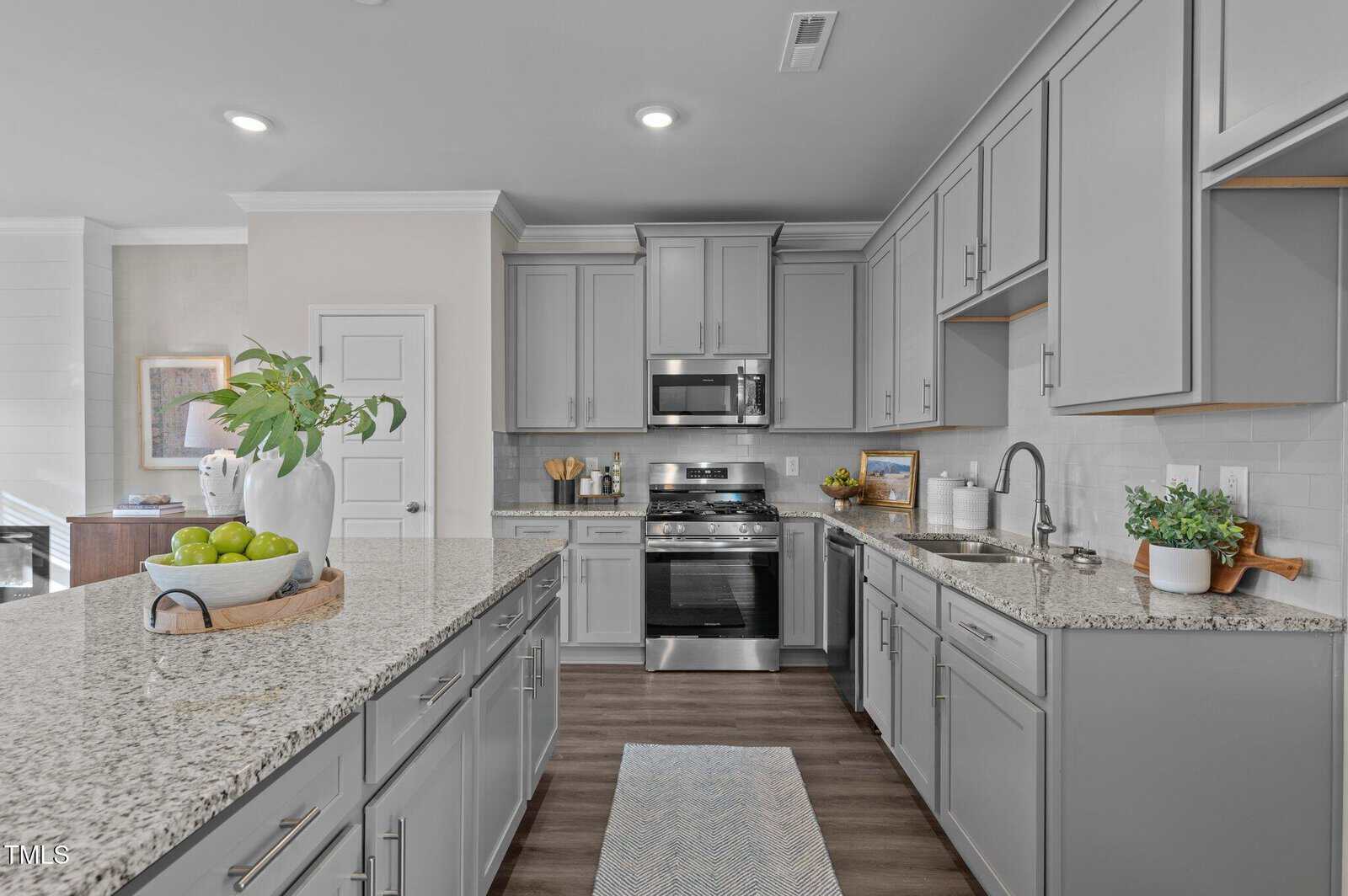 Modern kitchen with gray cabinets, stainless steel appliances, granite countertops, a kitchen island with a bowl of green apples, potted plants, and wooden flooring. Sink and stove are visible, with wall art and utensils on the counters.