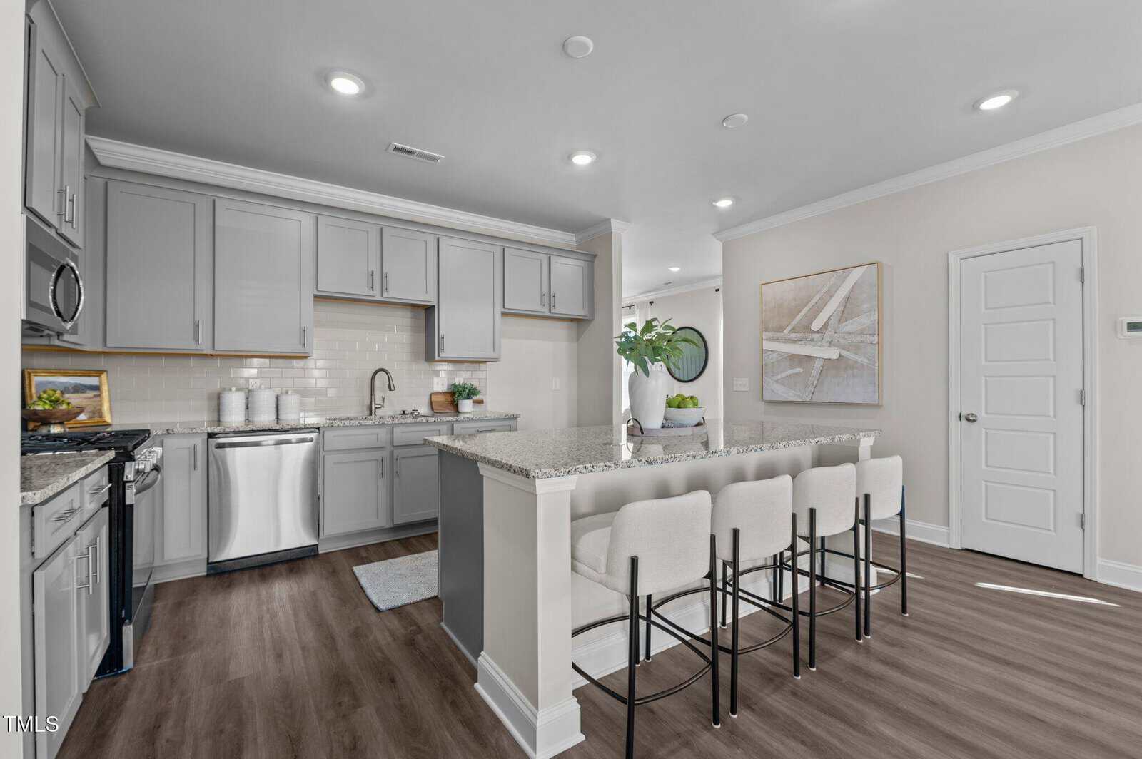 A modern kitchen with gray cabinets, stainless steel appliances, granite countertops, a white tile backsplash, and a large island with four upholstered bar stools. The space features wood flooring and neutral walls.