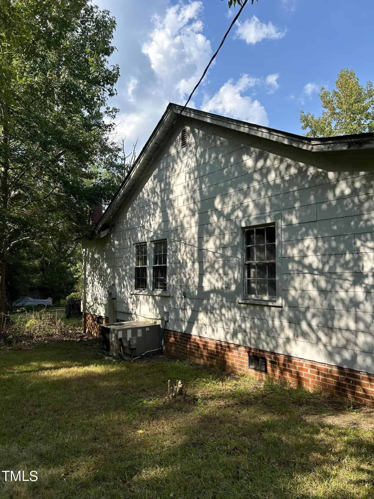 A rustic white house with a red brick foundation is partially shaded by trees. Sunlight filters through the leaves, casting intricate shadows on the exterior. The yard is overgrown with grass, and a utility unit is visible against the wall.