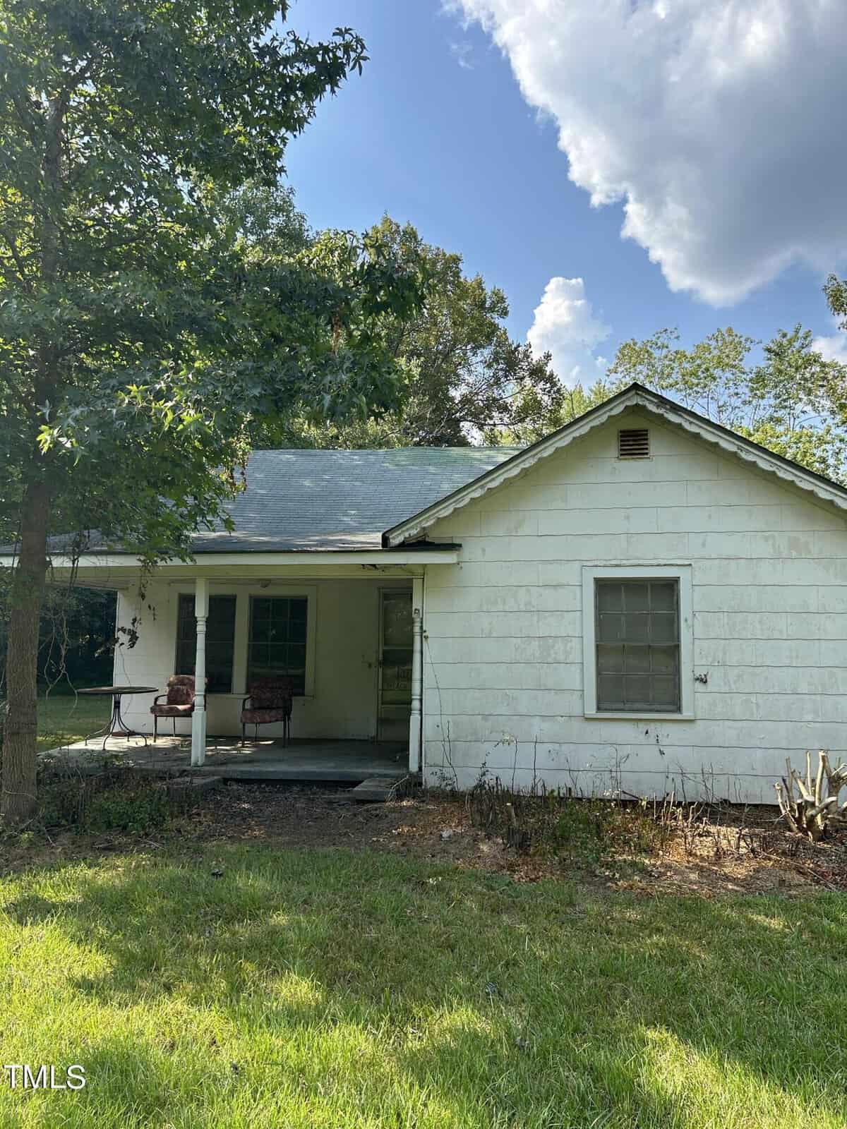 A small, weathered white house with a covered porch, a couple of chairs, and a green lawn. Surrounded by tall trees, the house is set under a partly cloudy sky. The overall appearance is rustic and peaceful.