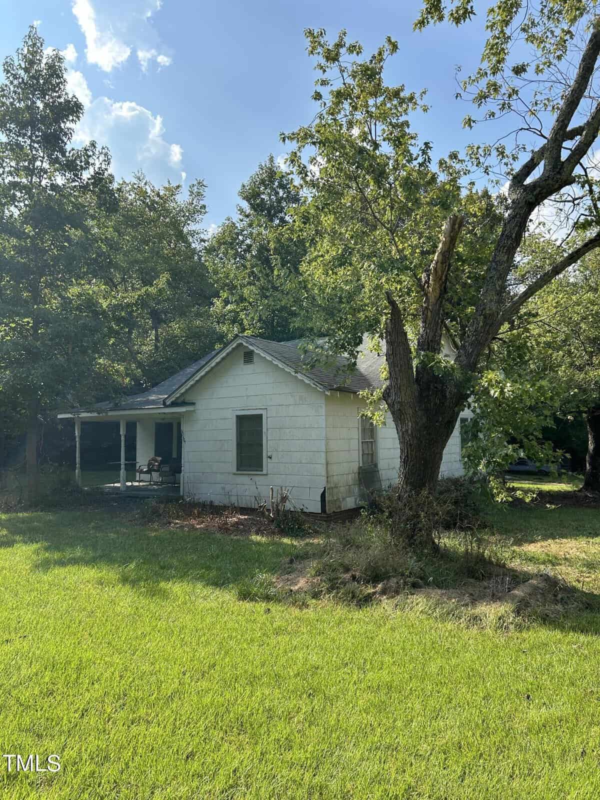 A small, weathered house stands in a grassy yard, partially shaded by tall trees. The white exterior and simple porch evoke a rustic, peaceful setting. Sunlight filters through the foliage, casting dappled shadows on the roof and surrounding lawn.