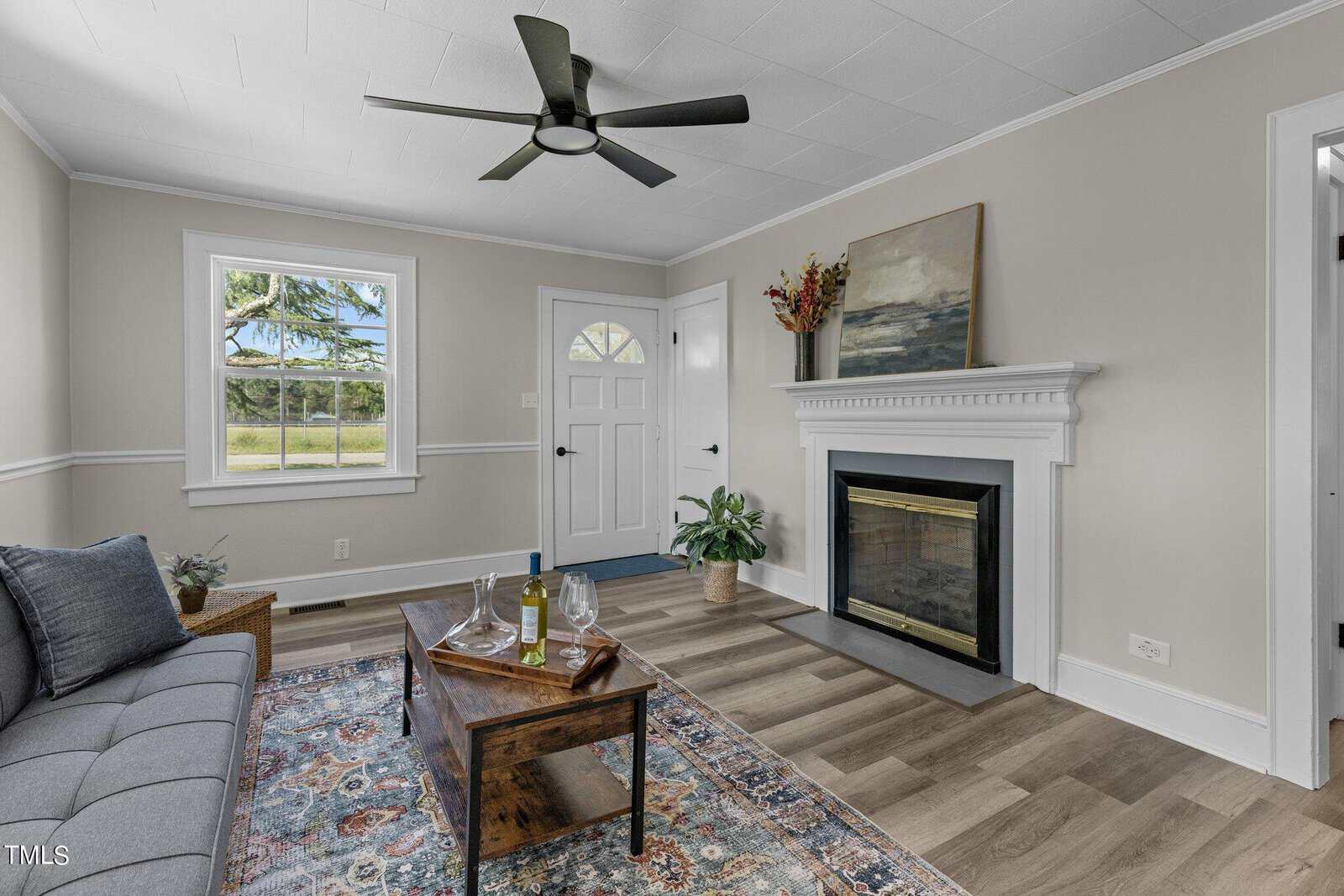 A cozy living room featuring a gray sofa with blue cushions, a wooden coffee table, and a fireplace with a white mantel. There is a painting above the fireplace and a potted plant beside it. The room has a window, door, ceiling fan, and an oriental rug on the floor.