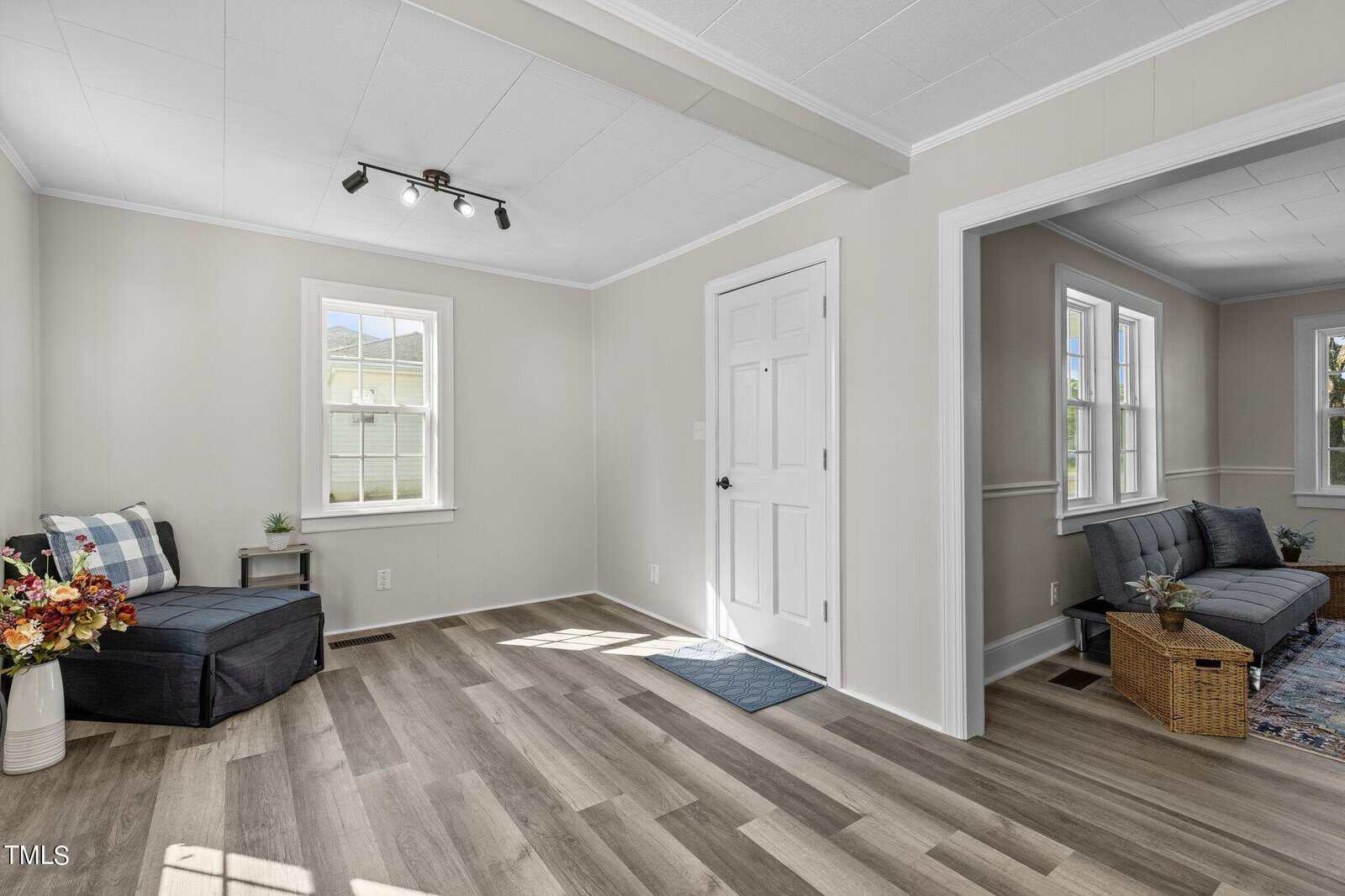 A bright room with light-colored walls and wood-paneled flooring. The space features a black armchair with a plaid throw pillow and blanket, a white door, and large windows letting in natural light. Another room with a rug and grey furniture is visible through an open doorway.