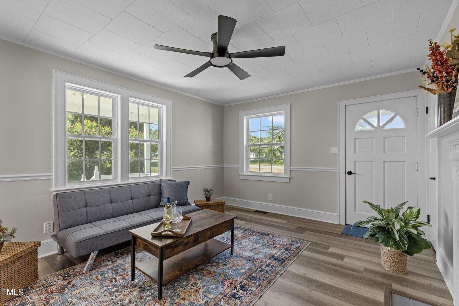 A cozy living room with light gray walls and wood flooring. It features a gray futon, a wooden coffee table, and a colorful patterned rug. A black ceiling fan is centered on the white ceiling, and there are two large windows and a white door letting in natural light.