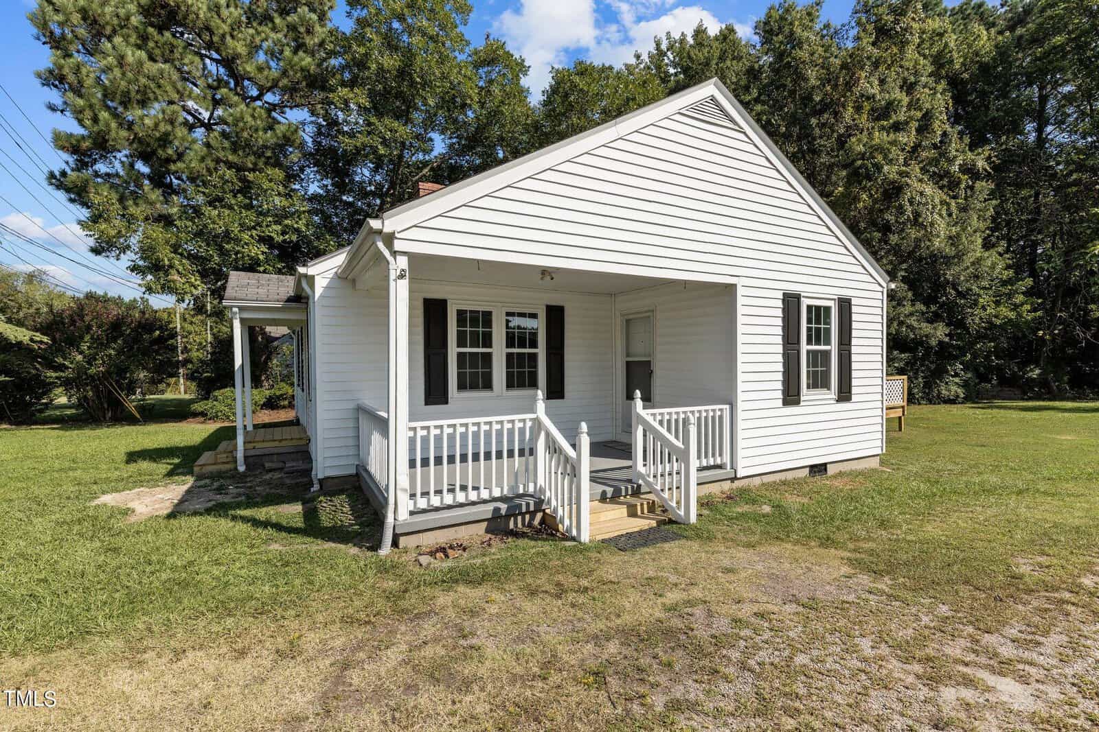 A small, white, single-story house with a covered front porch and white railings. The house has a modest grassy lawn and is surrounded by trees and greenery. The sky is blue with a few clouds.