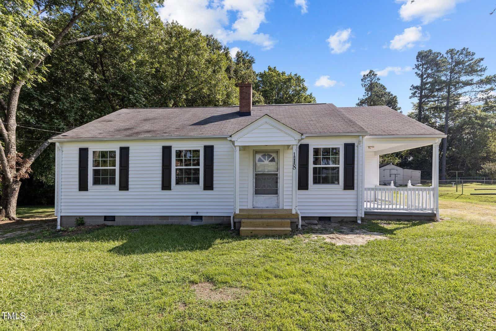 A white single-story house with black shutters and a gable roof sits at 118 Spring Valley Drive, Henderson. The home features a small front porch with steps leading up to the door, set on a grassy lawn with trees in the background under a bright blue sky with scattered clouds.