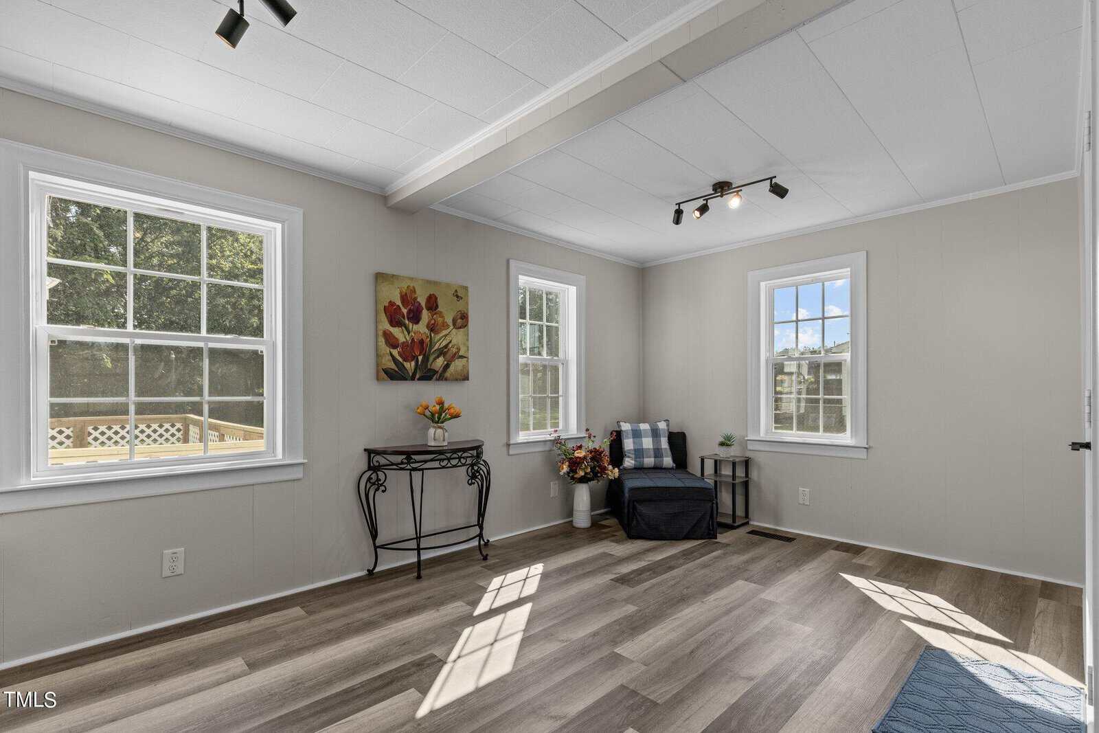 A bright room with white walls and two large windows allowing natural light in. There are wood-style floors, a blue armchair with a checkered pillow, a small black metal table with flowers, and a red flower painting on the wall.