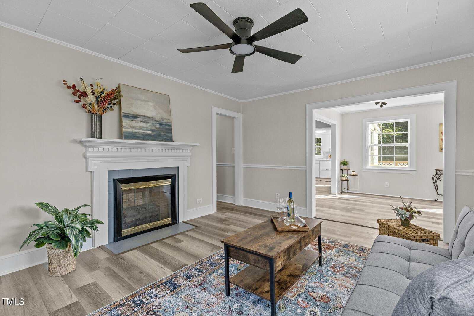 A cozy living room with light-colored walls and wood flooring. It features a white fireplace with a plant on the left side and a painting above it. A gray couch and wooden coffee table are centered on a colorful rug. A ceiling fan is above, and a doorway leads to a bright kitchen.