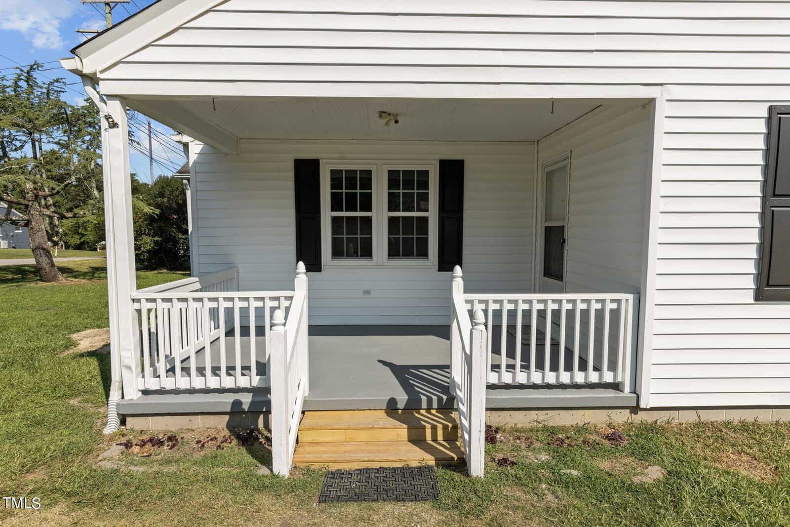 A small white wooden porch with black shutters and white railing attached to a house. The porch has a covered roof, a wooden step, a railing with vertical bars, and an outdoor light mounted on the ceiling. The surrounding yard has green grass and trees.