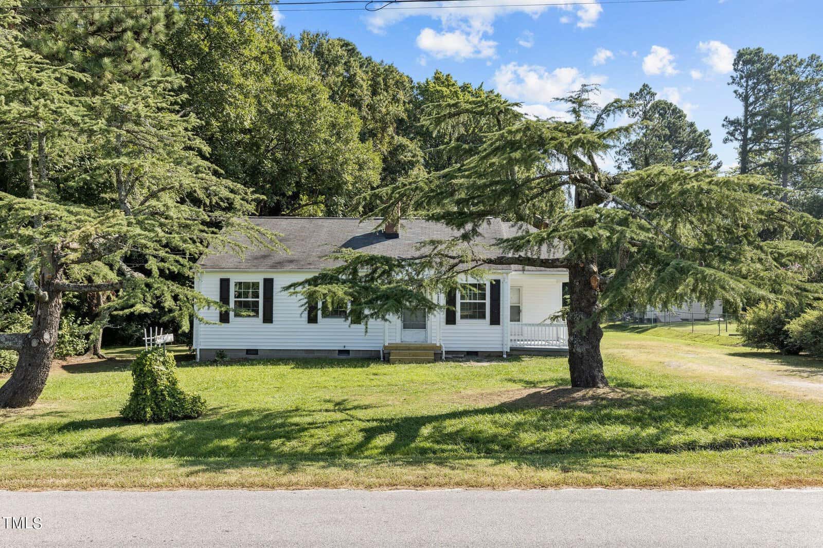 A small white house with a covered front porch sits on a grassy lawn, bordered by tall evergreen trees. A paved road runs in front of the property, and the sky above is clear with a few scattered clouds.
