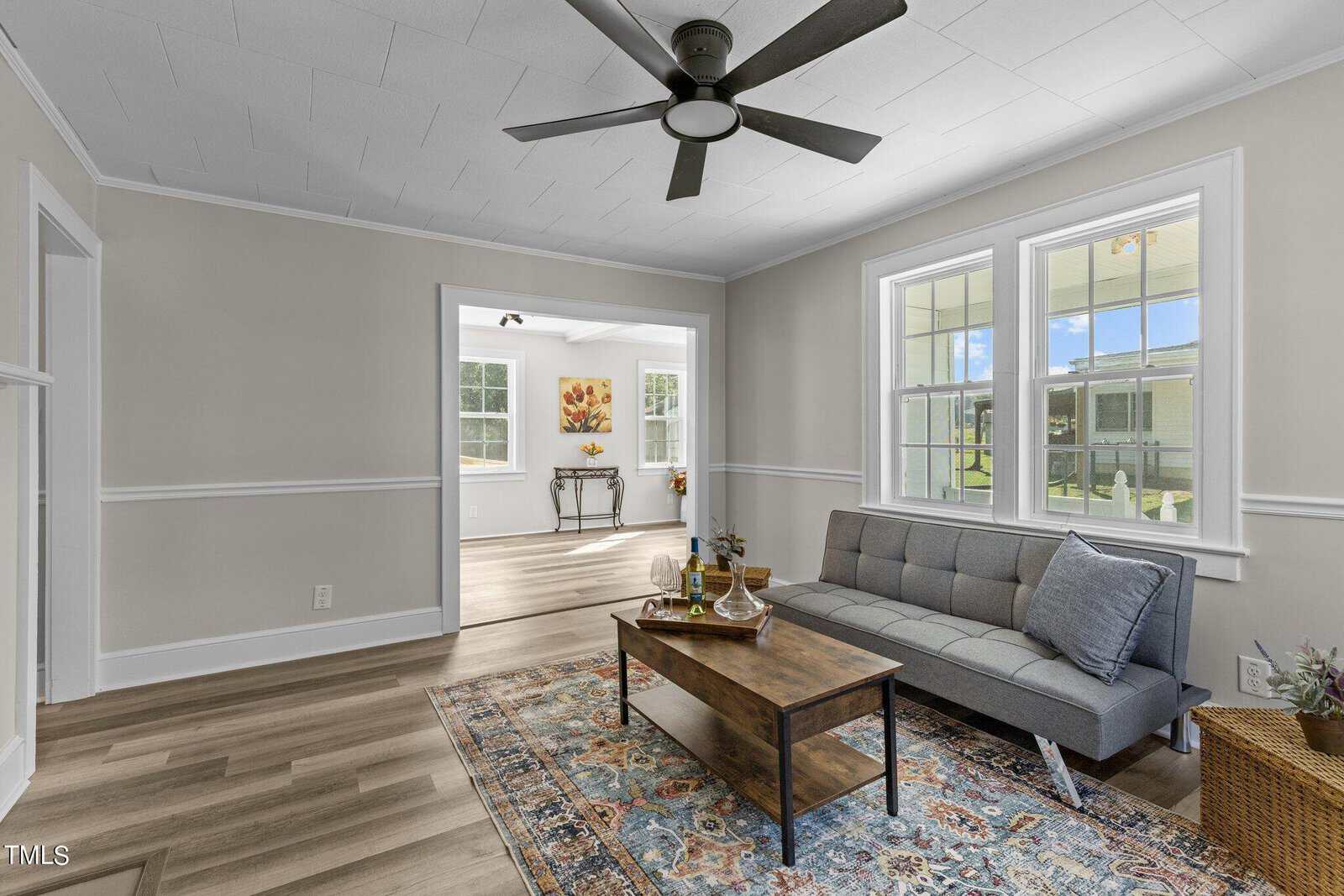 A bright living room featuring a gray futon sofa, a wooden coffee table, and a patterned rug. Large windows let in natural light. A ceiling fan is overhead, and an adjacent room with more windows and a table is visible in the background.