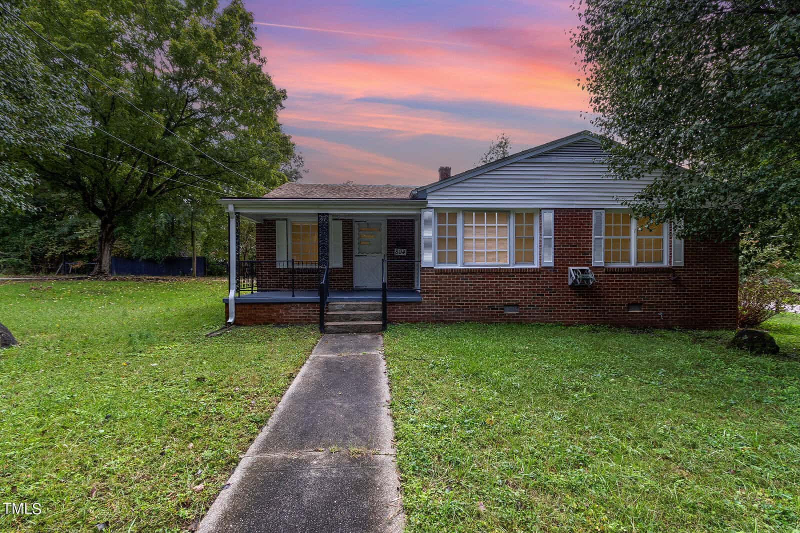 Located at 804 Eastside Drive, this charming single-story brick house in Henderson features a welcoming porch and steps leading to the front door. The yard boasts lush grass, trees, and a concrete path. The sky is alive with pink and orange hues, signaling a serene sunset or sunrise.