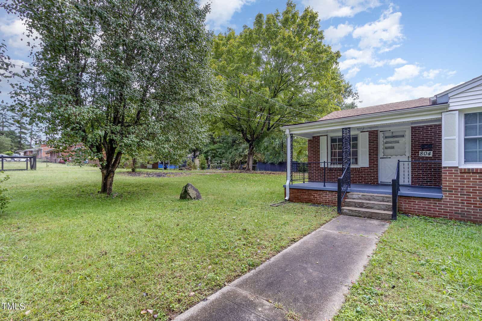 A small brick house with a front porch and stairs, surrounded by a grassy yard with large trees. The sky is partly cloudy, and a narrow path leads to the porch. The house has white trim and a visible house number "604.