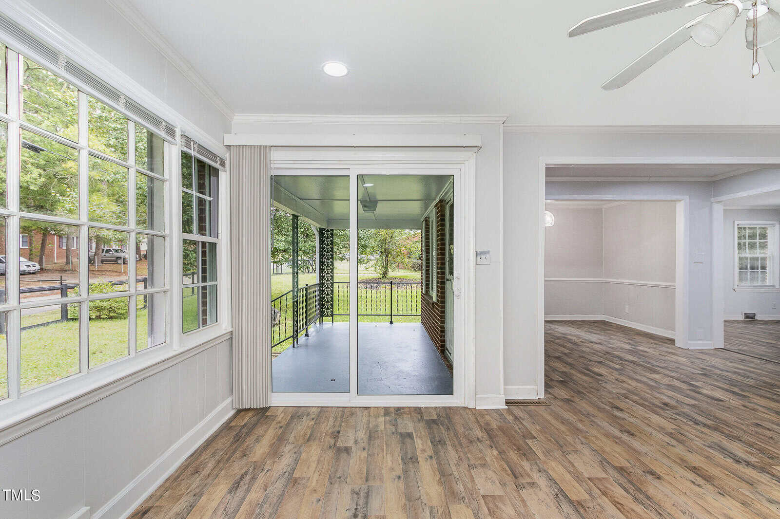 A bright, open room with large windows on the left, sliding glass doors leading to an outdoor area in the center, and an open doorway to another room on the right. The room has light-colored walls, wood flooring, and a ceiling fan.