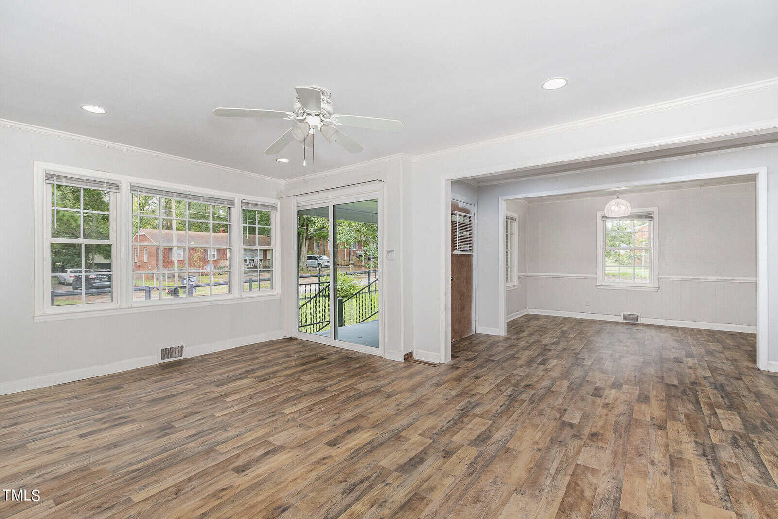 Spacious living area with light wood flooring, ceiling fan, large windows, and sliding doors leading to a porch. Adjacent room with similar flooring and a chandelier. White walls and molding add a modern, airy feel.