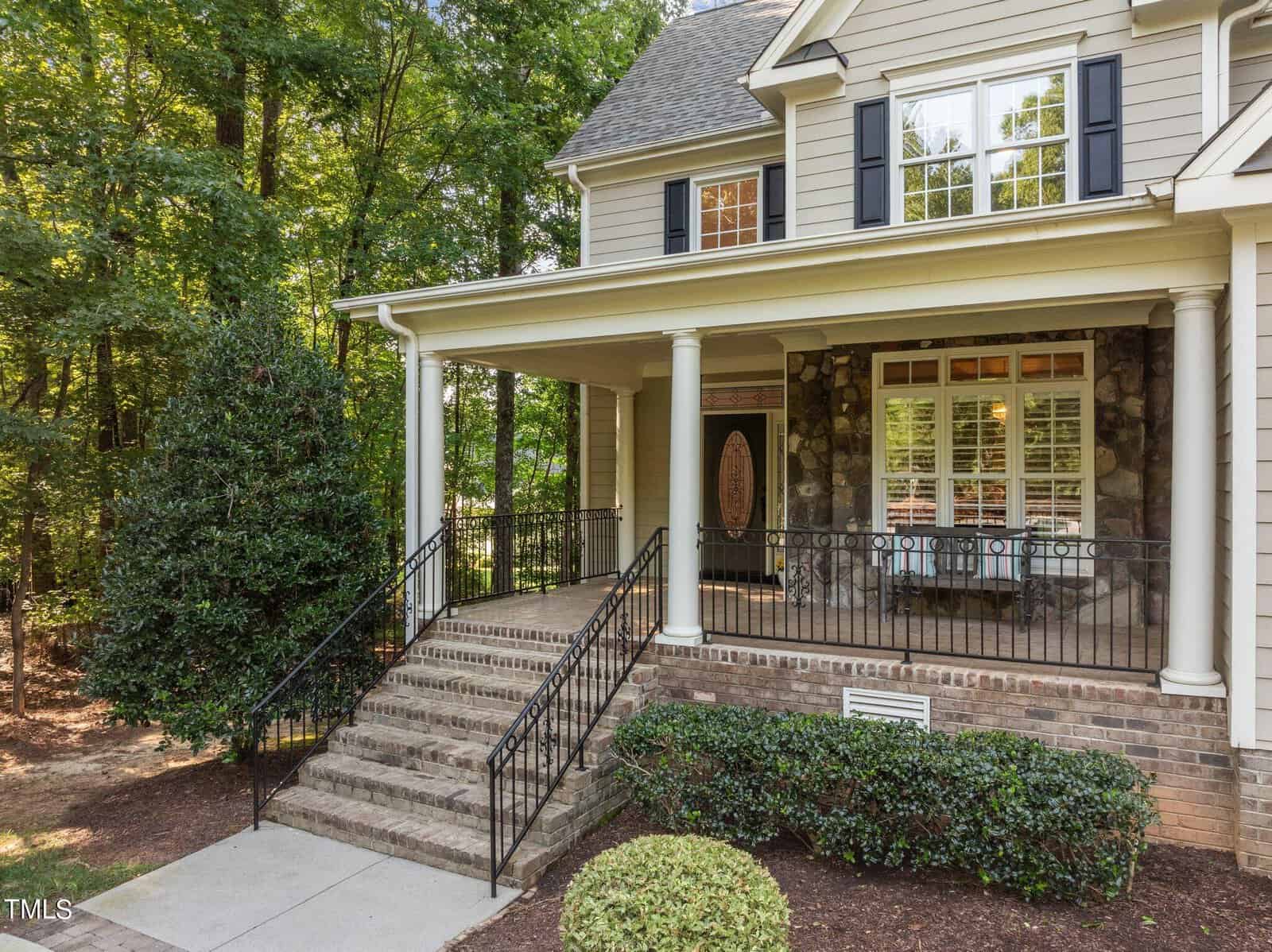A two-story house with a stone facade features a covered front porch and a set of brick steps leading to a wooden door with decorative glass. The porch is supported by white columns and is surrounded by bushes and trees.