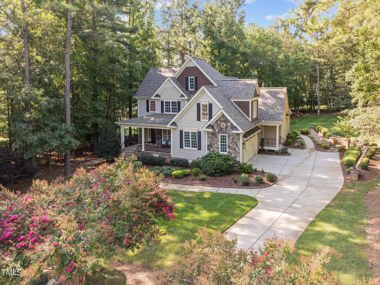 A two-story house with gray siding and a large front porch is surrounded by trees. The property features a landscaped garden, a curved driveway, and a well-maintained lawn.