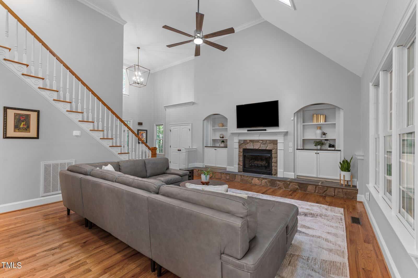 Spacious living room with high ceilings, featuring a large gray sectional sofa, a stone fireplace, a mounted TV, and built-in shelves. Wooden floor and staircase, with a ceiling fan and natural light from large windows. Minimalist decor in neutral tones.
