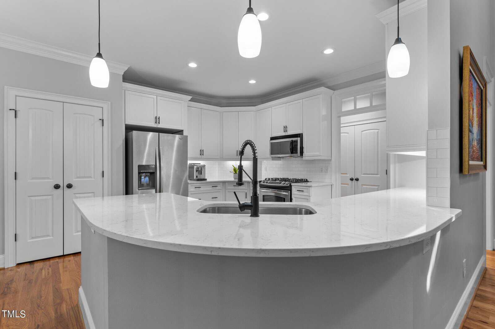 A modern kitchen with white cabinets and a marble countertop island. It features a stainless steel refrigerator, built-in microwave, and a gas stove. Pendant lights hang from the ceiling, and there's a single vase with flowers on the counter.