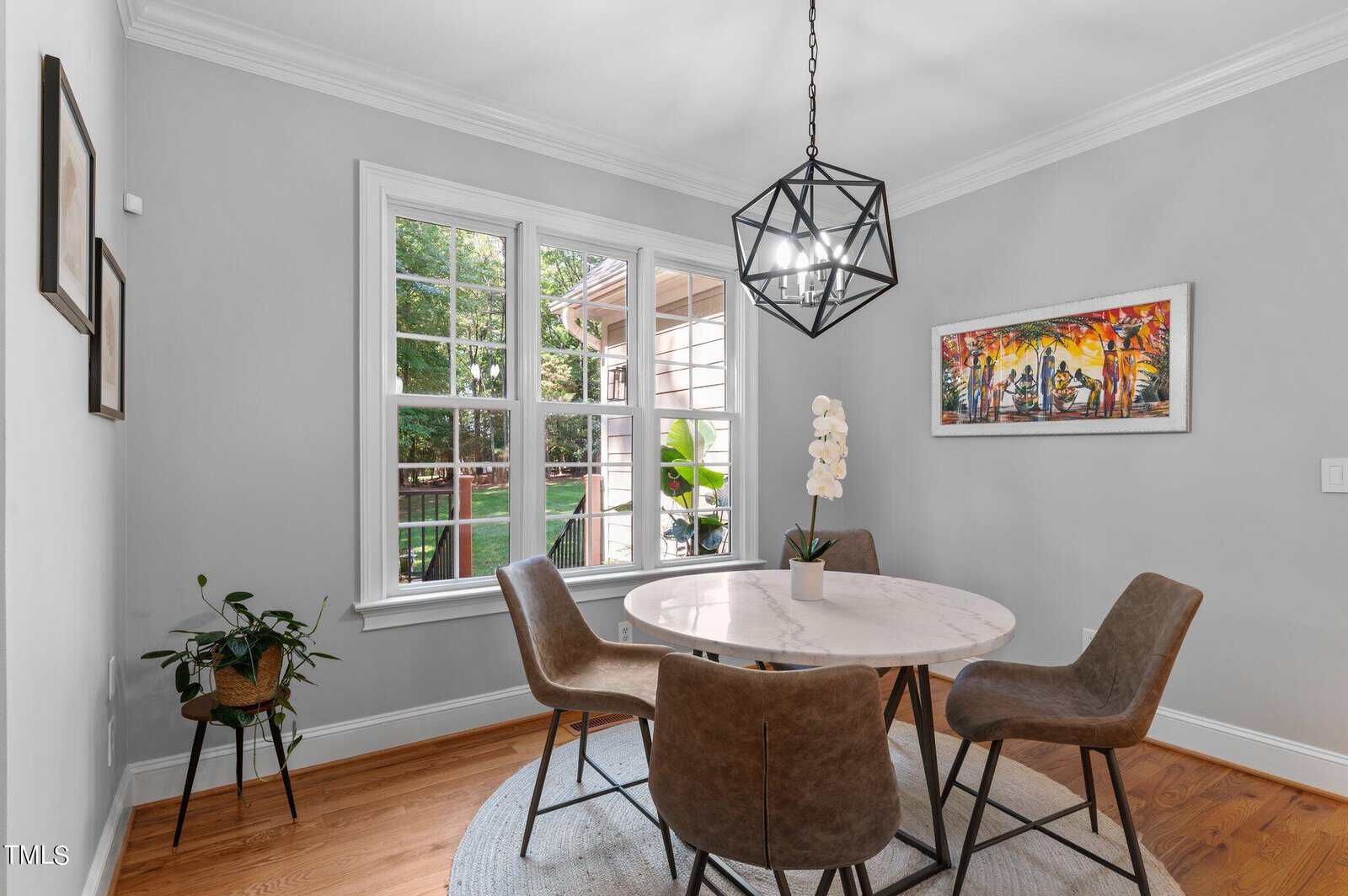 A dining area with a round marble table surrounded by four brown chairs. A geometric pendant light hangs above. A potted plant sits in the corner, and a colorful painting is on the wall. Large windows reveal a view of the outdoors.