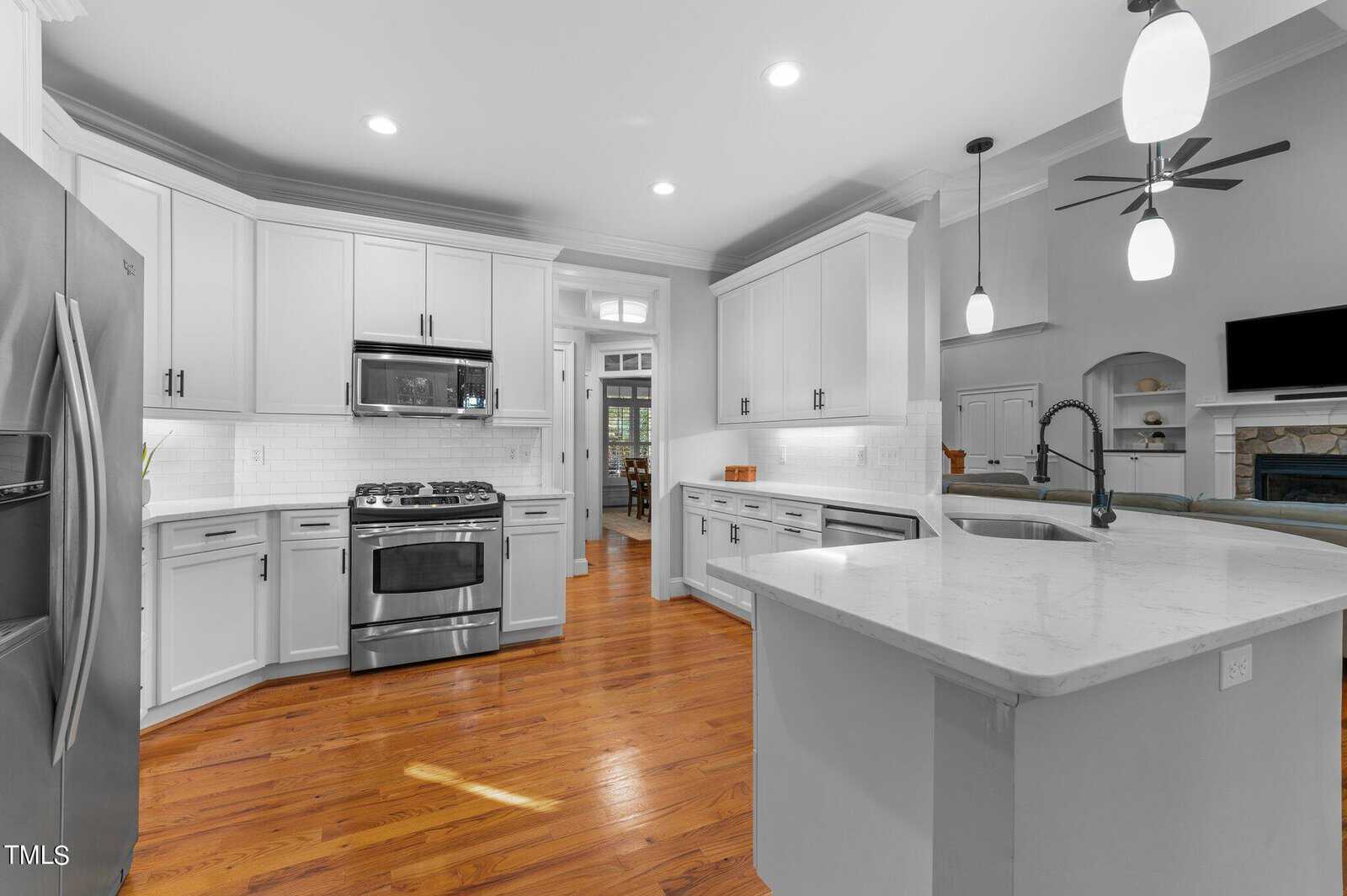 A modern kitchen with white cabinets and stainless steel appliances, including a refrigerator and stove. The room features wooden flooring and an island with a sink. Pendant lights hang from the ceiling. An open doorway leads to another room.