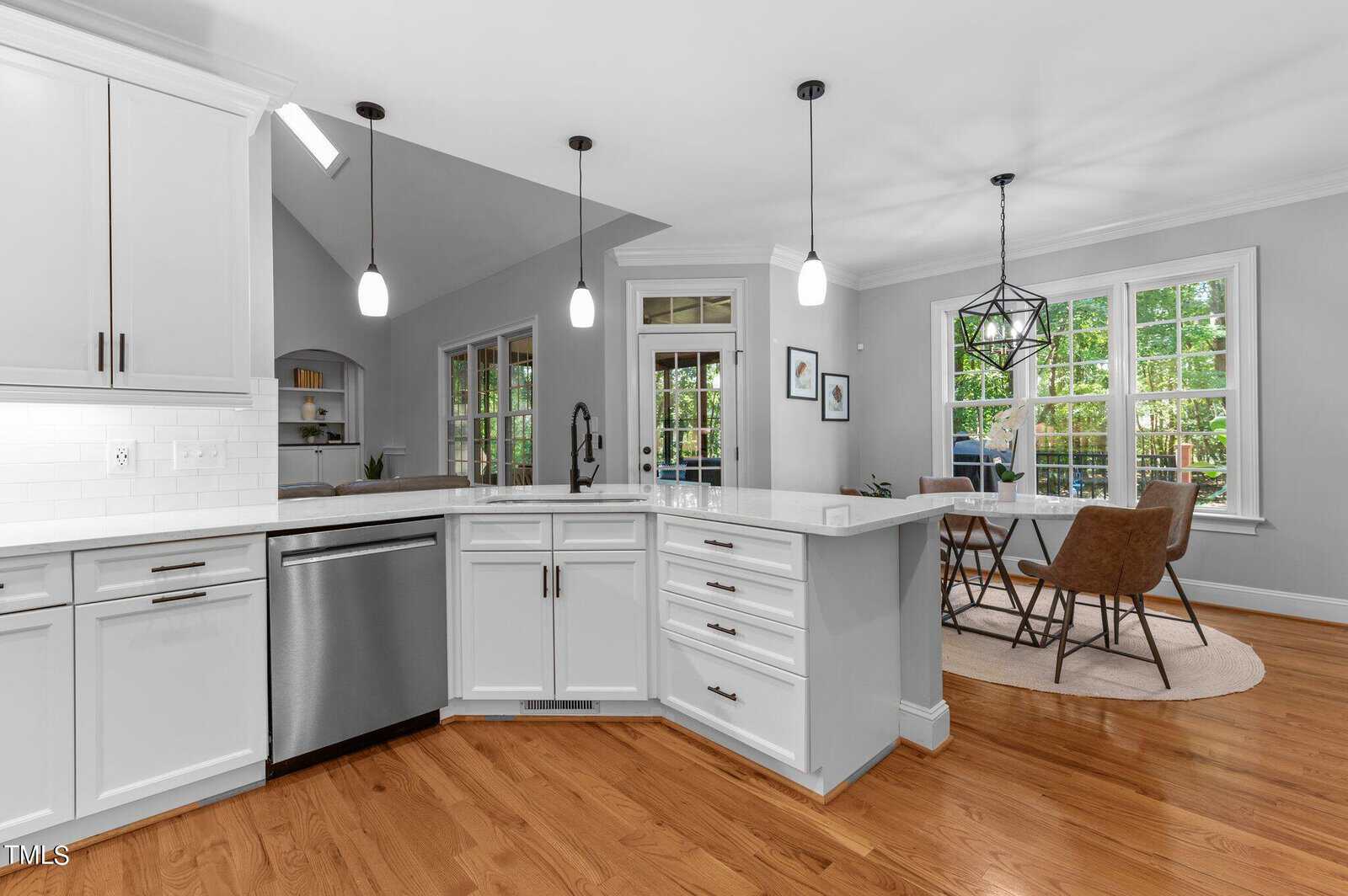 A modern kitchen with white cabinets and a stainless steel dishwasher. Three pendant lights hang over the island. The open dining area features a round table with four brown chairs. Large windows provide natural light, and wooden floors are visible.