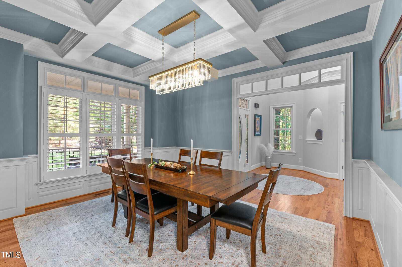 A dining room with a wooden table and six chairs. A modern chandelier hangs above the table, which has three candles on it. The room has light blue walls, a coffered ceiling, and wood flooring. Large windows and a doorway are visible.