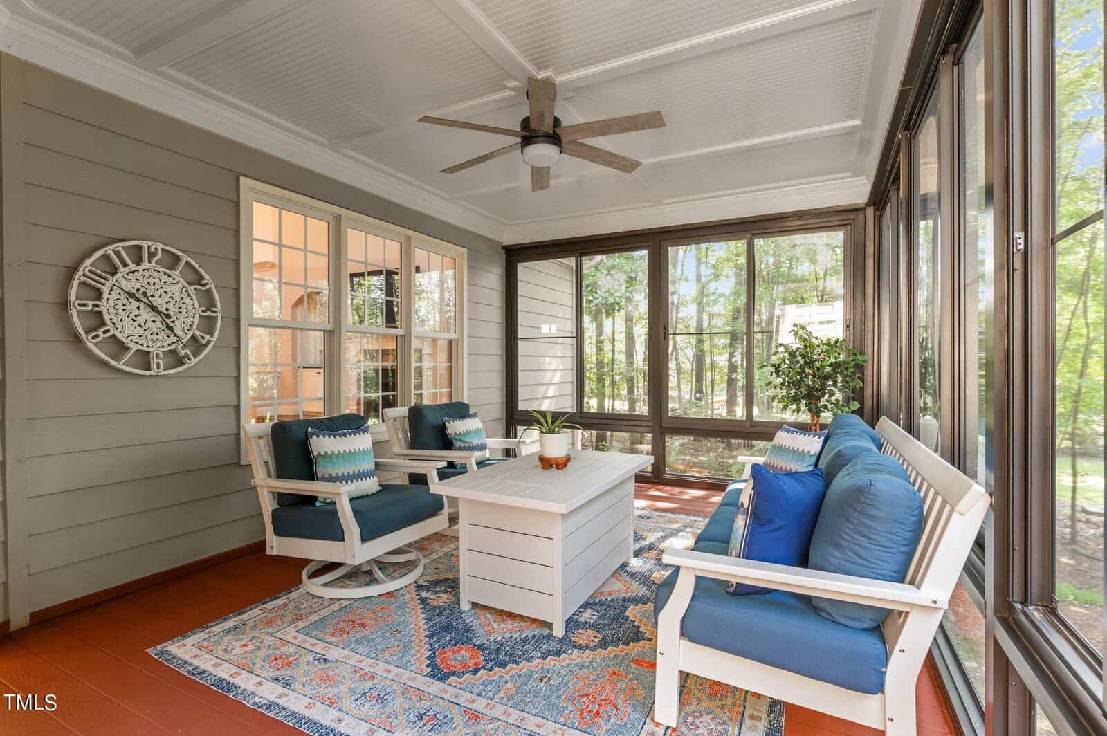 A sunroom with a white ceiling fan, glass windows, and a seating area. The furniture includes a white sofa and chairs with blue cushions around a white coffee table. A decorative clock is on the wall, and an indoor plant is in the corner.