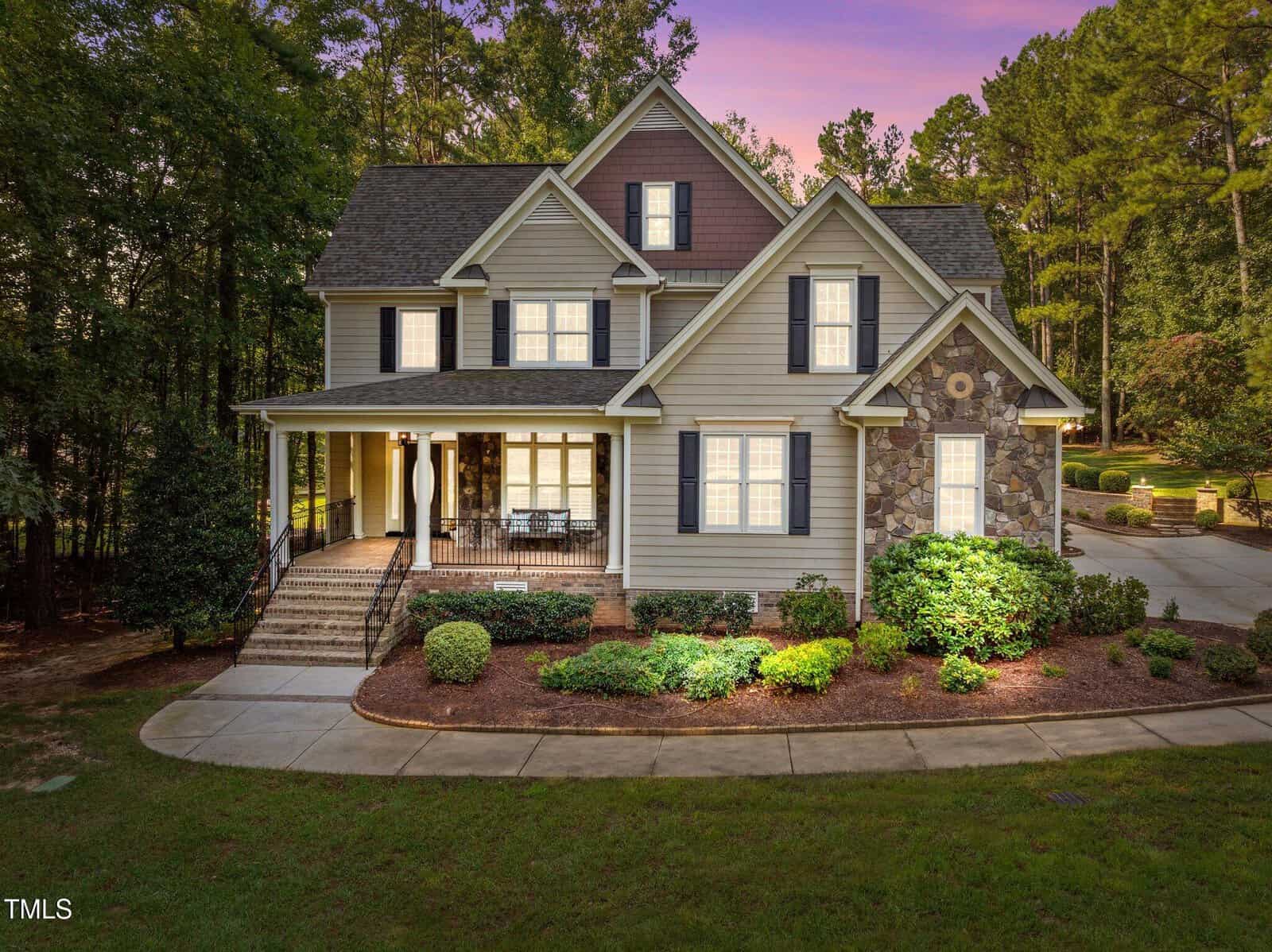 The charming two-story house at 4011 Cashmere Lane in Youngsville boasts a mix of siding and stone exterior, complemented by a spacious front porch and numerous windows. A curving driveway winds through meticulously landscaped greenery, under a sky tinted with the hues of sunset or twilight.
