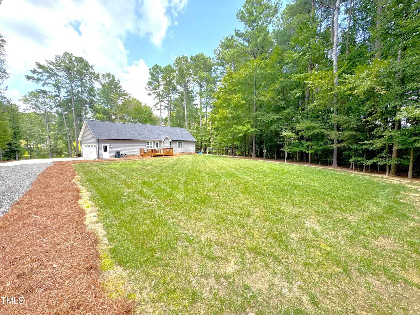 A single-story house with a gray roof sits in the background, surrounded by pine trees. It has a long driveway and a large green lawn in the foreground under a partly cloudy sky.