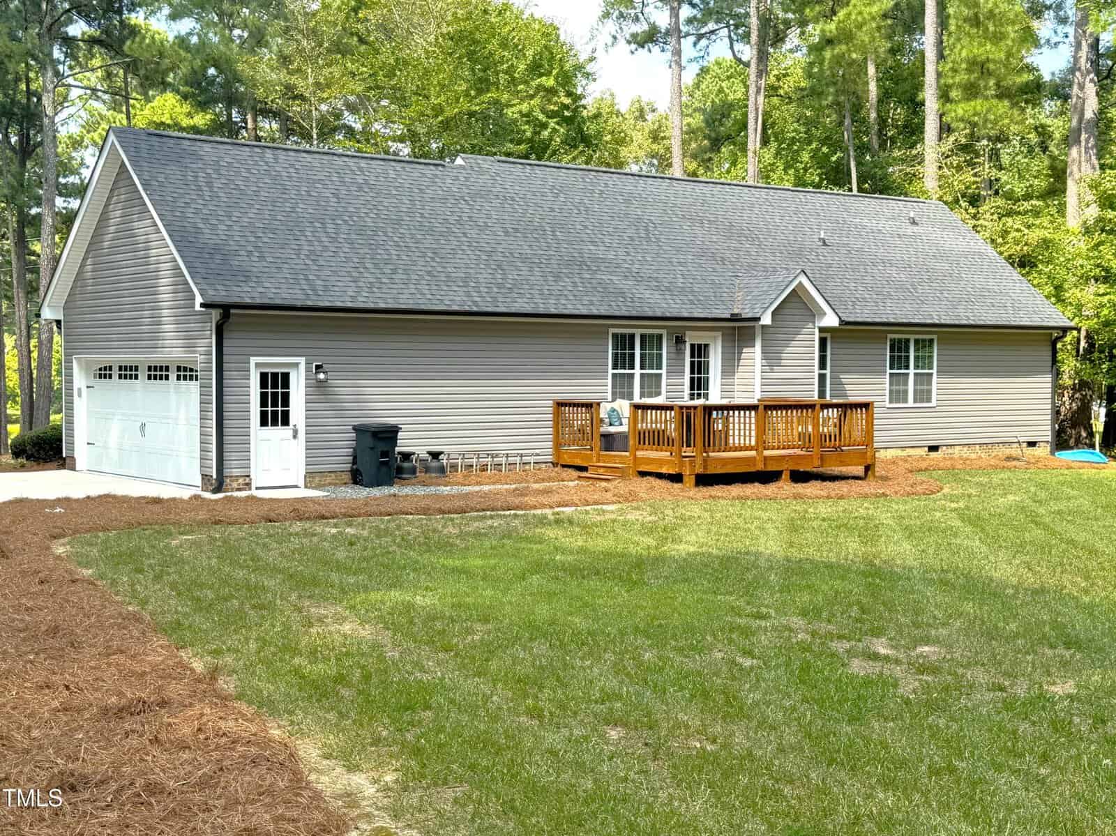 A single-story gray house with a black roof is set in a lush green yard. It features a wooden deck on the side and an attached garage. Tall trees surround the property, creating a serene forest backdrop.