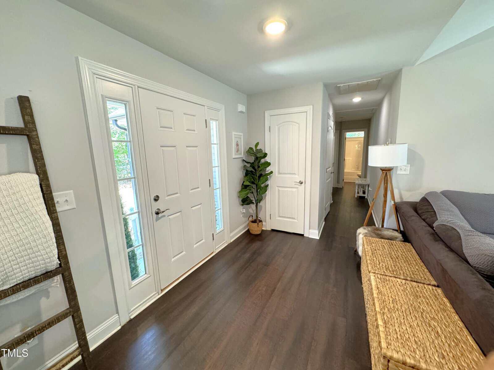 A modern entryway with a glass-paneled front door, a potted plant, and a beige sofa. The walls are light gray and the floor is dark wood. A basket and a wooden ladder with a white blanket enhance the decor.