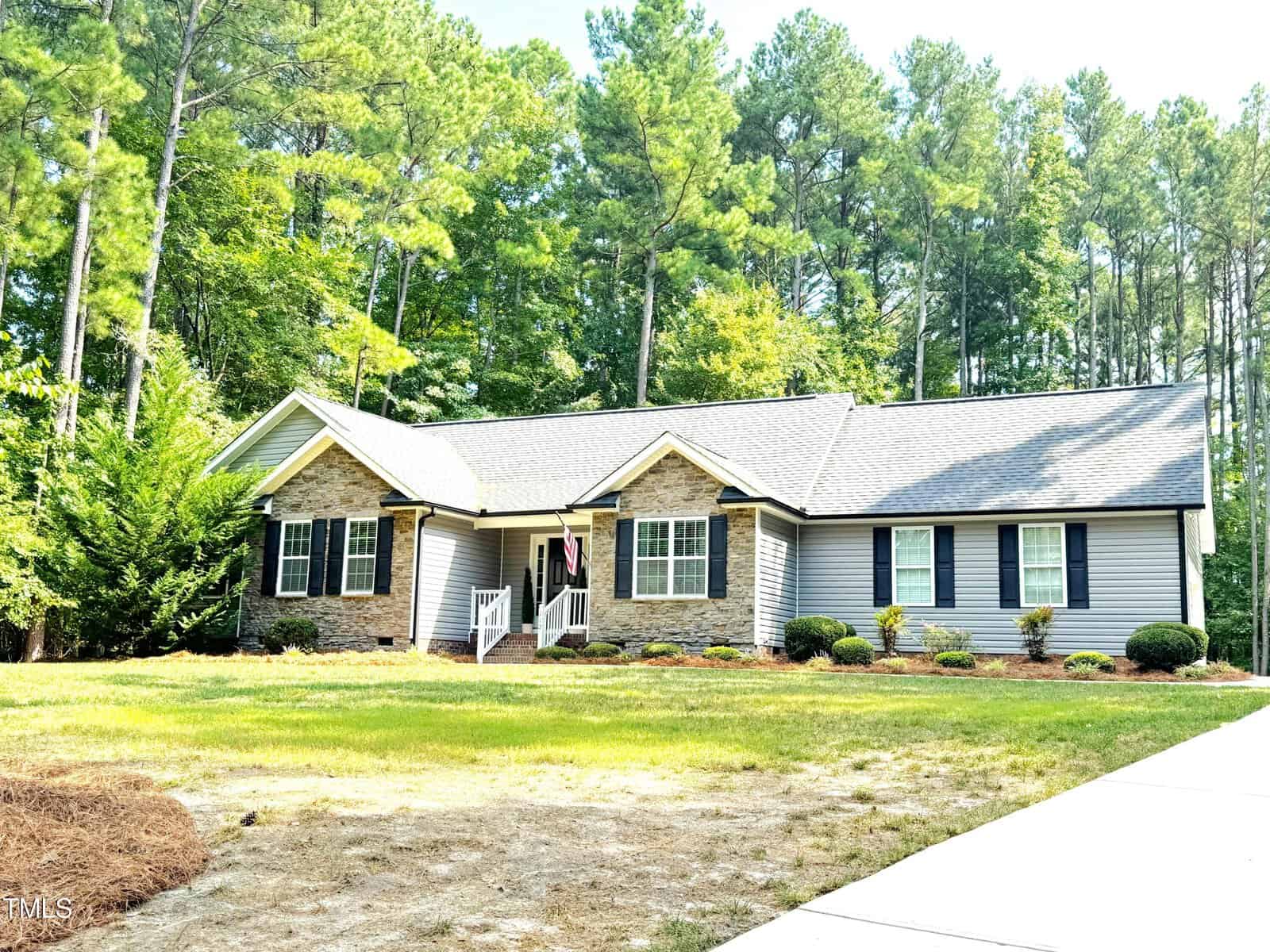 Nestled at 112 Franklin Street, this single-story house features gray siding and stone accents. A covered front porch with an American flag is set against tall trees, surrounded by neat landscaping and a spacious lawn. A driveway gracefully leads to the inviting home.