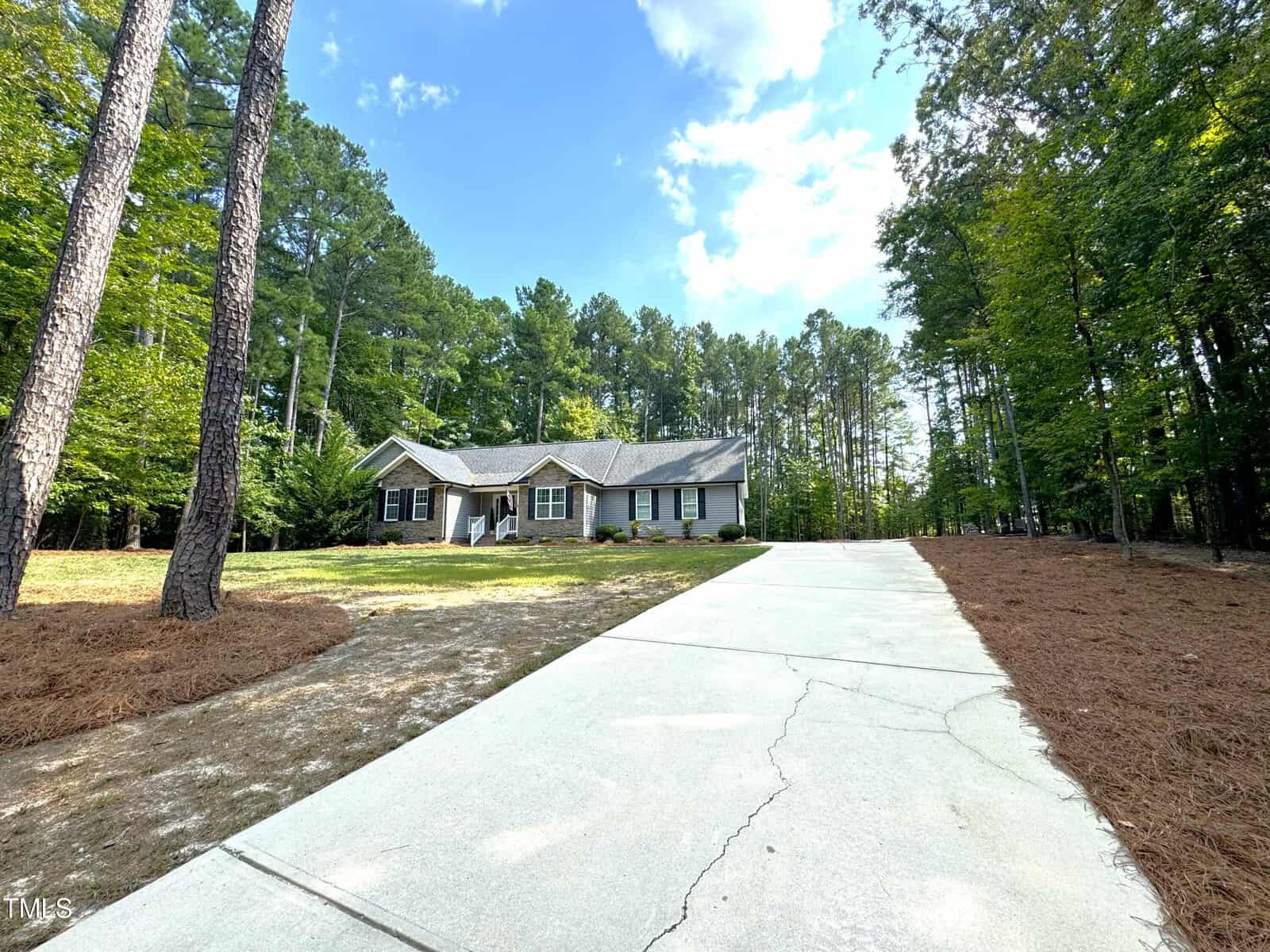 A single-story house with a gray exterior is surrounded by tall trees. A long cement driveway leads to the house, bordered by grassy and pine needle-covered areas. The sky is partly cloudy with patches of blue visible.