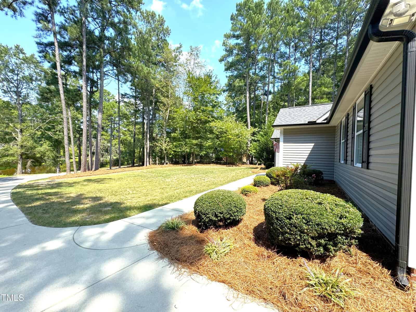 A house with light siding and a gray roof is surrounded by a manicured lawn and shrubs. A curved driveway leads to the house, and tall trees line the background under a clear blue sky.