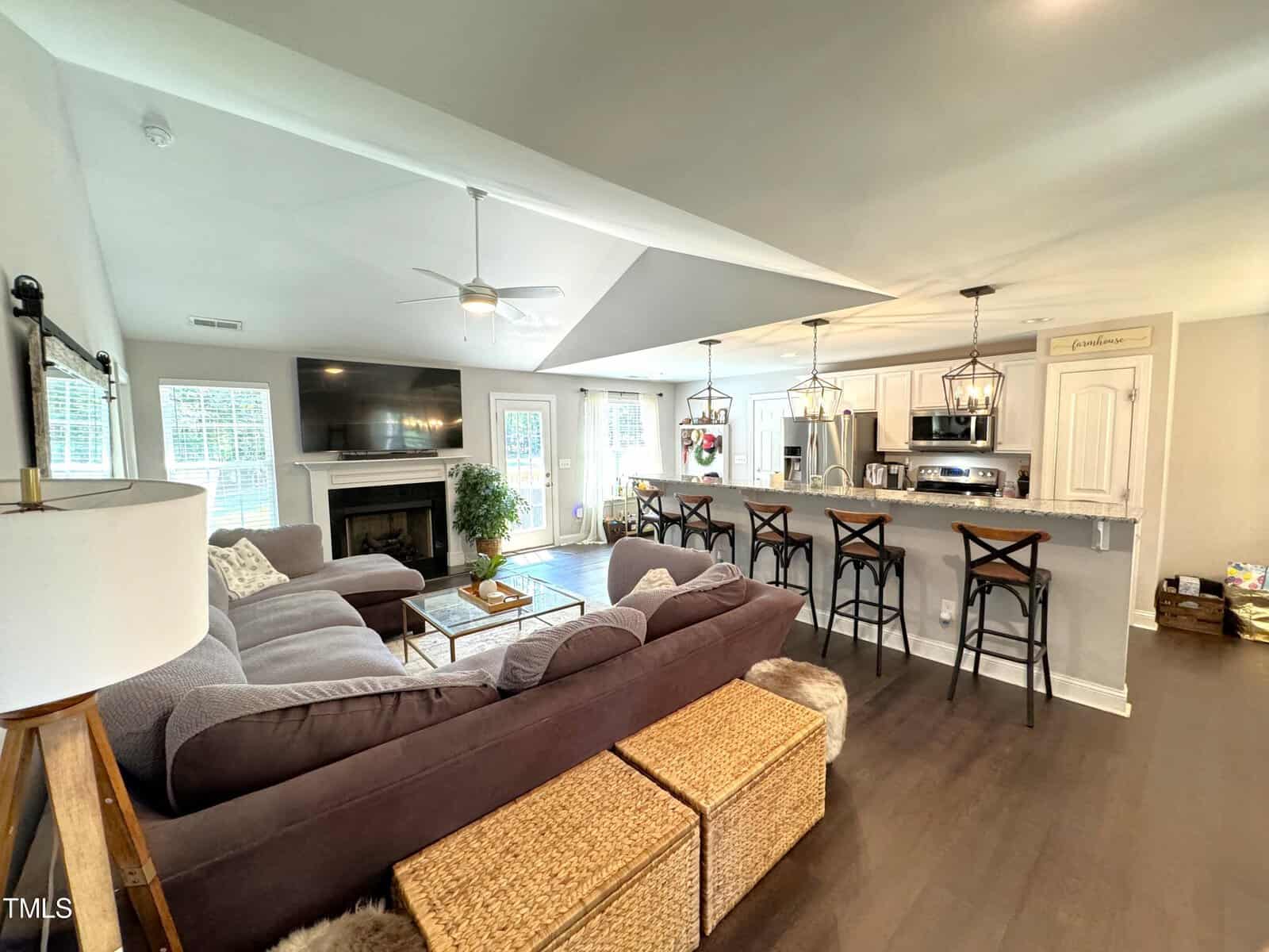 A spacious living room with a large L-shaped gray sofa and a fireplace. The open kitchen features a bar counter with four stools. Natural light streams in through multiple windows, highlighting the wooden floor and modern decor.