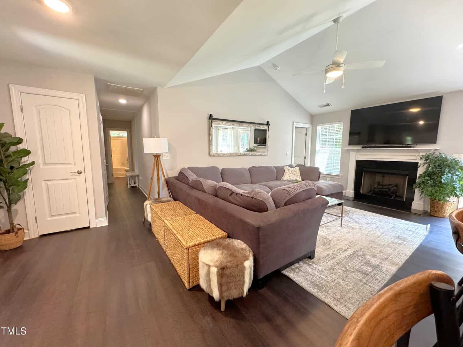 A cozy living room with a brown sectional sofa, a large TV above a fireplace, and a woven basket table. Soft lighting illuminates the space, showcasing a potted plant and a stylish floor lamp. The room has a vaulted ceiling and wooden flooring.