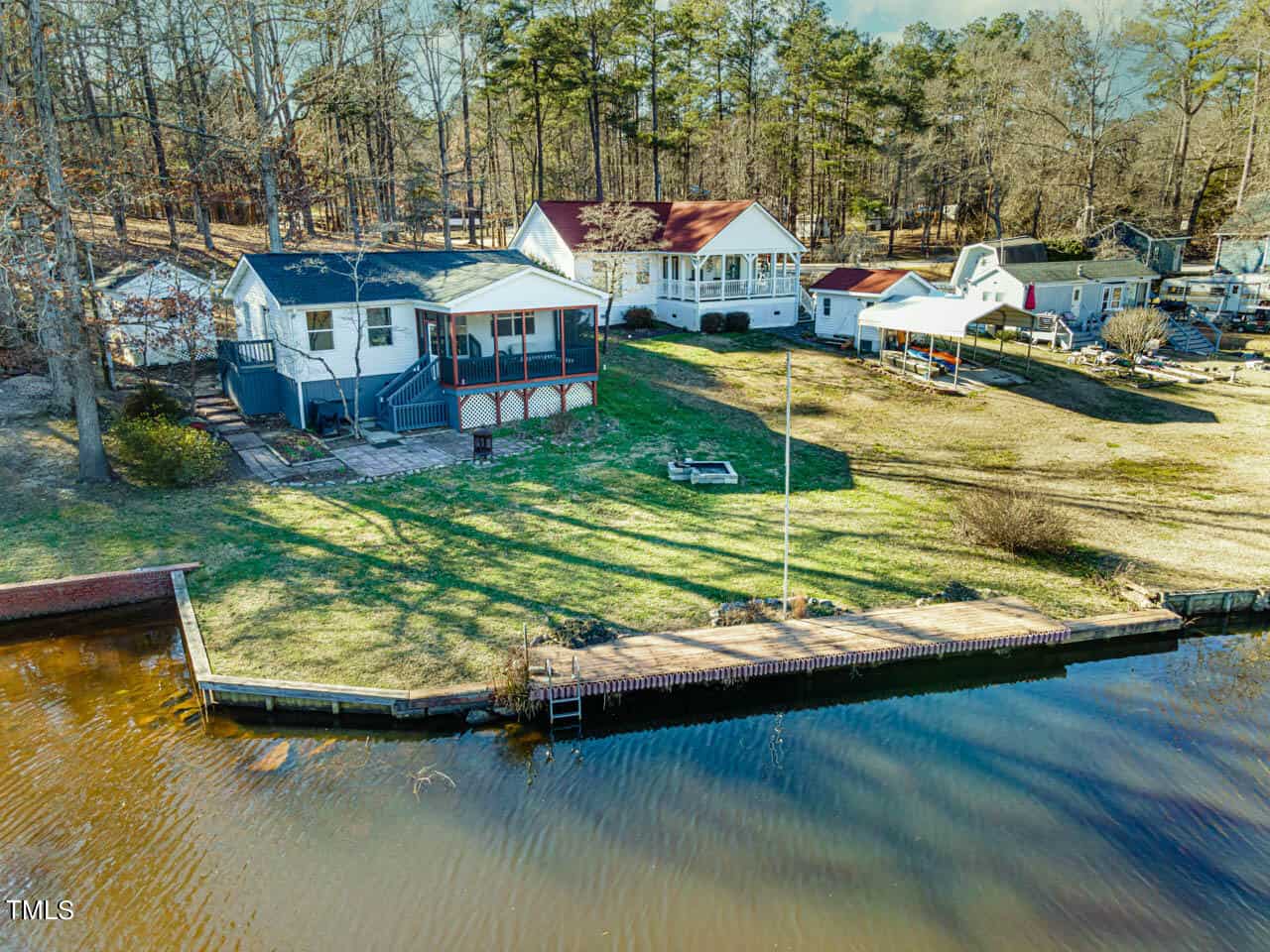 Aerial view of lakeside homes with docks, grassy yards, and trees in the background. The shoreline features a wooden retaining wall and a small pier extending over the water.