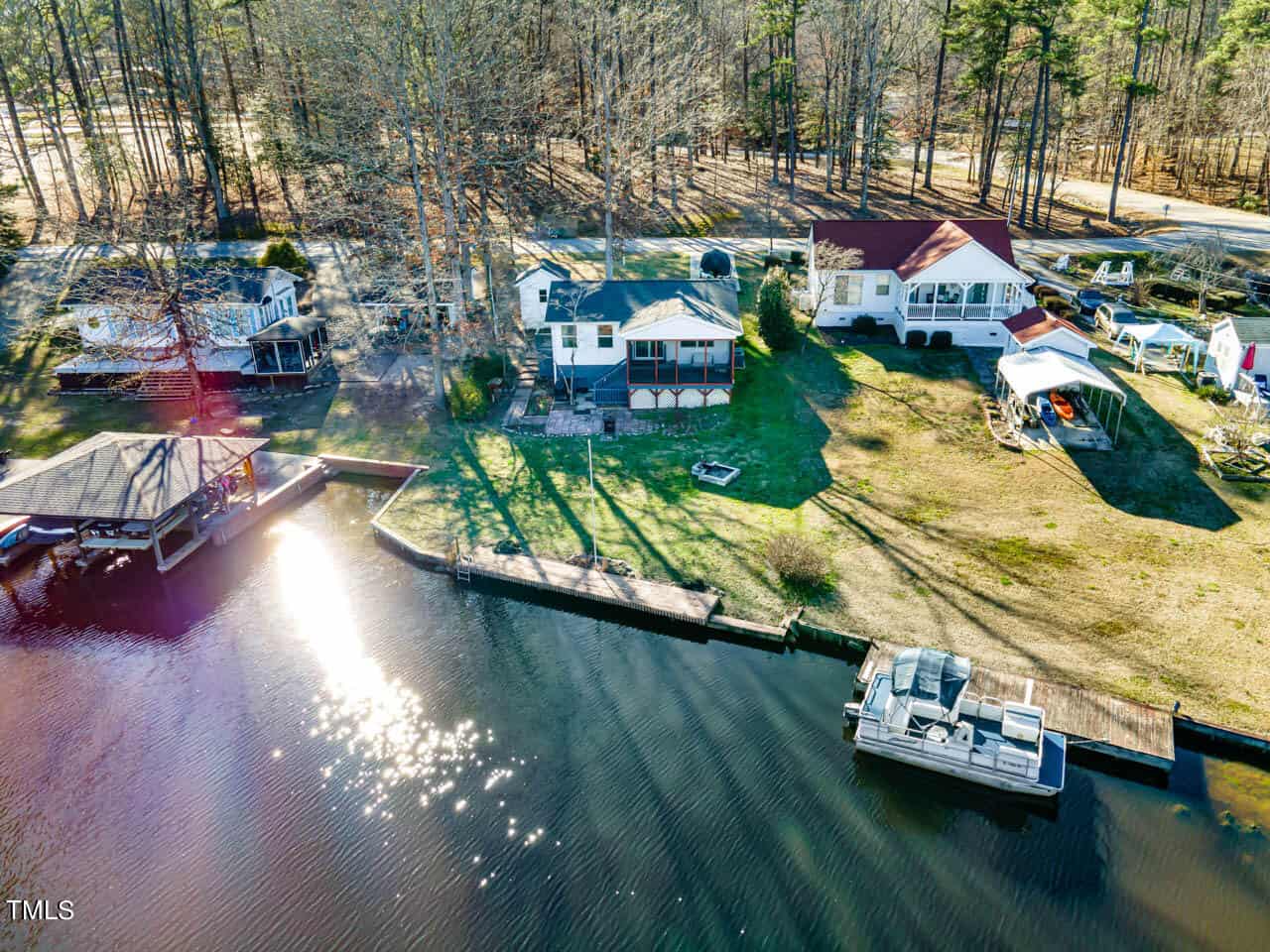 Aerial view of three houses by a wooded area next to a lake with a docked boat, covered patio, and a gazebo structure on the water. Sunlight reflects off the water and the trees cast long shadows on the grass.