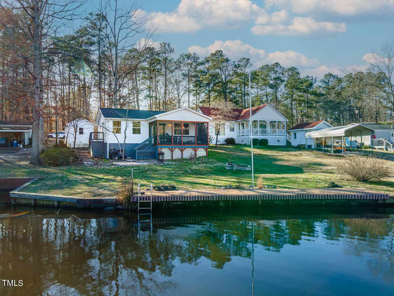 A group of white houses with porches and docks sits on a grassy, tree-lined shoreline beside a calm body of water under a partly cloudy sky. A small ladder leads from the water to the grassy bank.