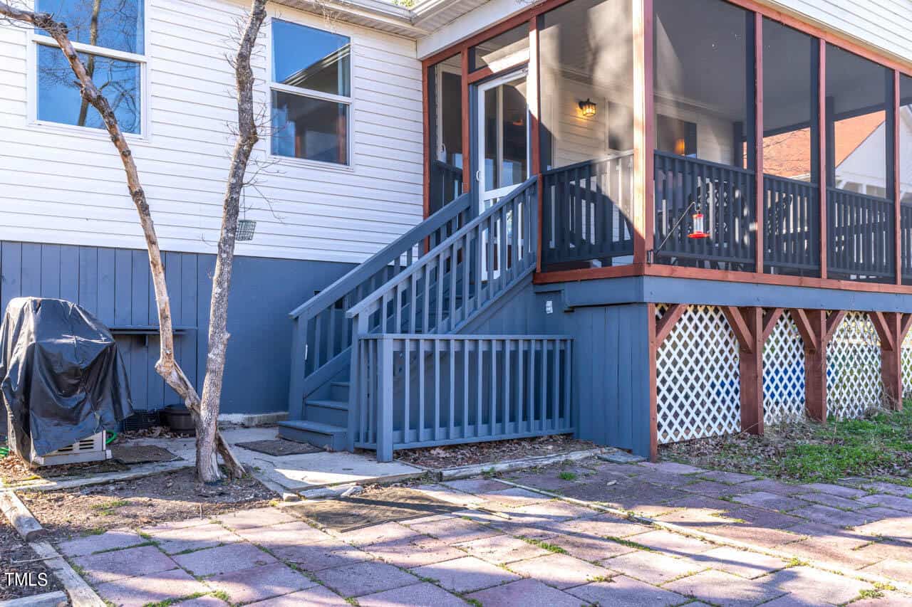 A backyard view showing a small staircase with a handrail leading to a screened porch attached to a house. There is a barbecue grill covered nearby and a paved patio area in the foreground.