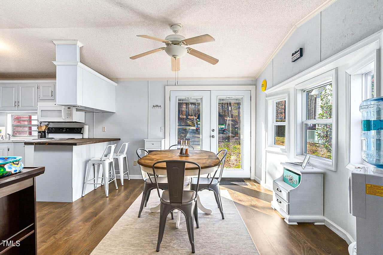 Bright kitchen with white cabinets, a round wooden dining table with four chairs, French doors leading outside, and large windows. A ceiling fan and a water dispenser are also visible. Sunlight streams in, illuminating the room.