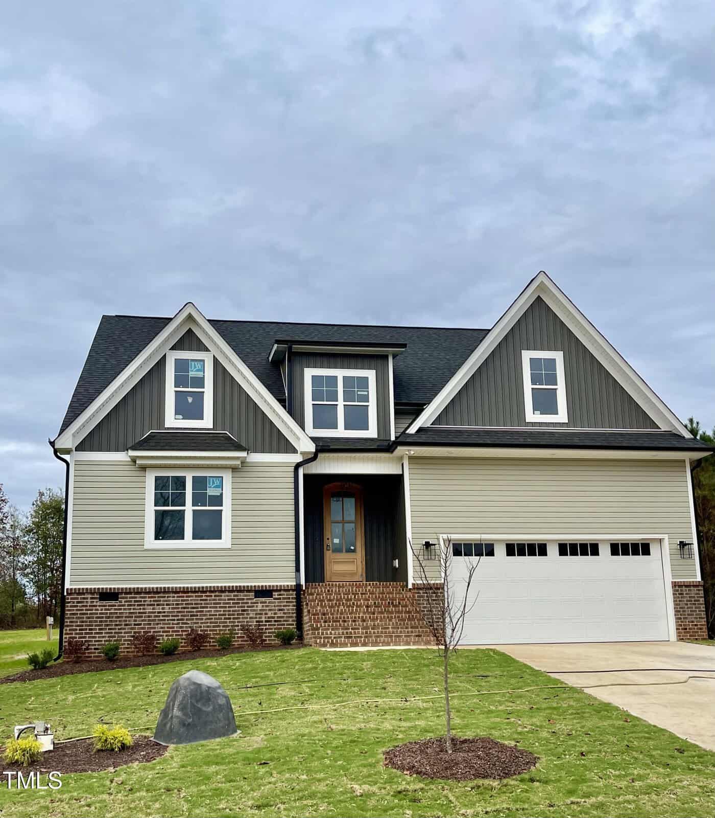 A modern two-story house with gray siding, brick accents, and a black roof. It has a double garage, wooden front door, and newly planted grass and landscape features in the front yard under a cloudy sky.