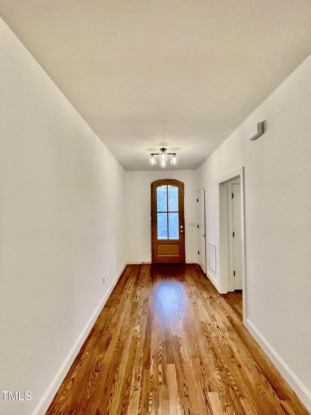 A narrow hallway with white walls, wooden flooring, and a wooden door with a window at the end. There are two open doorways on the right side and a ceiling light fixture near the door.