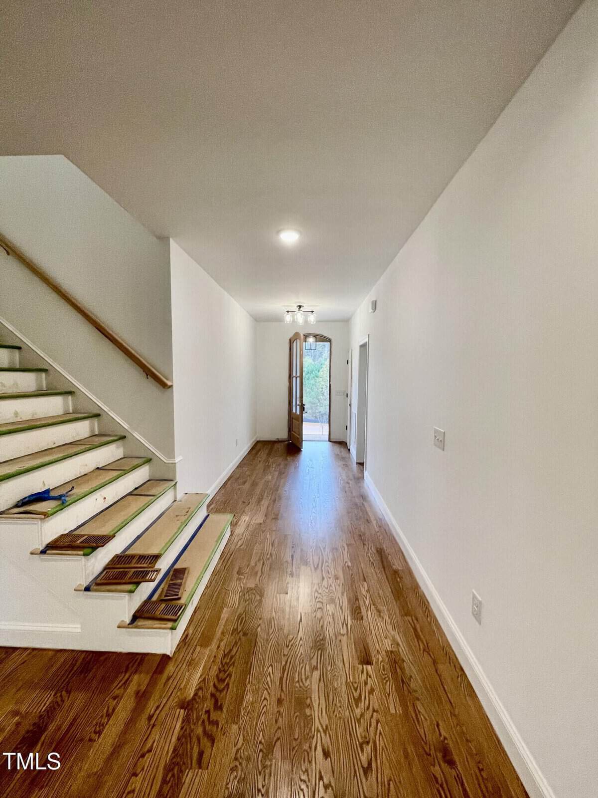 A hallway with hardwood floors and white walls, an unfinished staircase on the left, and an open wooden front door at the far end letting in natural light.