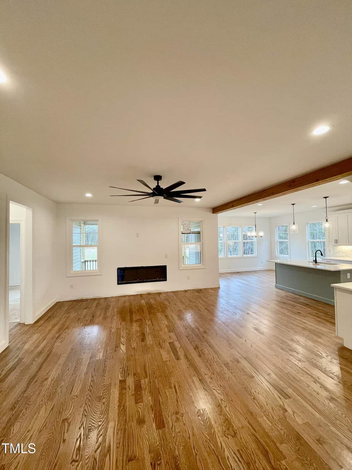 Spacious open-concept living area with light wood floors, a black ceiling fan, modern fireplace, and large windows. The room extends to a bright kitchen with an island and pendant lights.