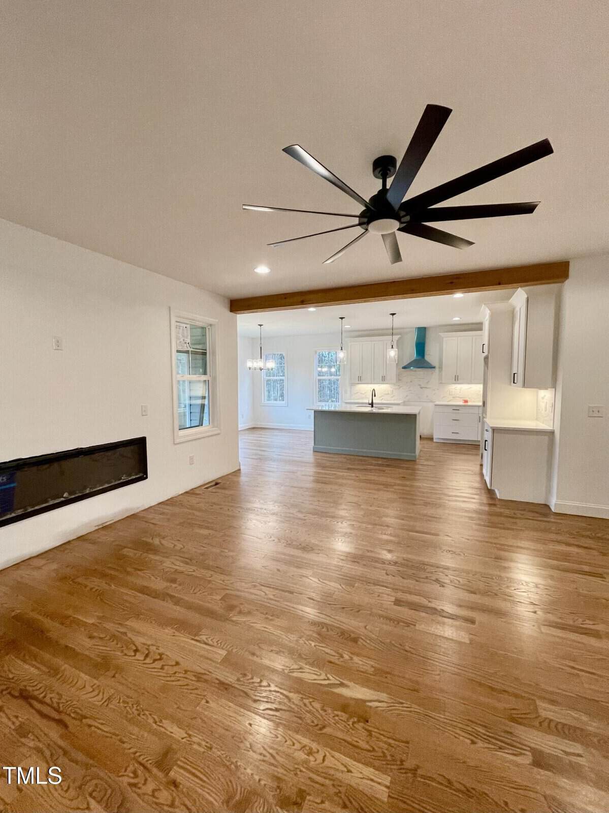 Open-concept living area with hardwood floors, a modern ceiling fan, a wall-mounted fireplace, and a spacious kitchen featuring an island, white cabinets, and a teal range hood.